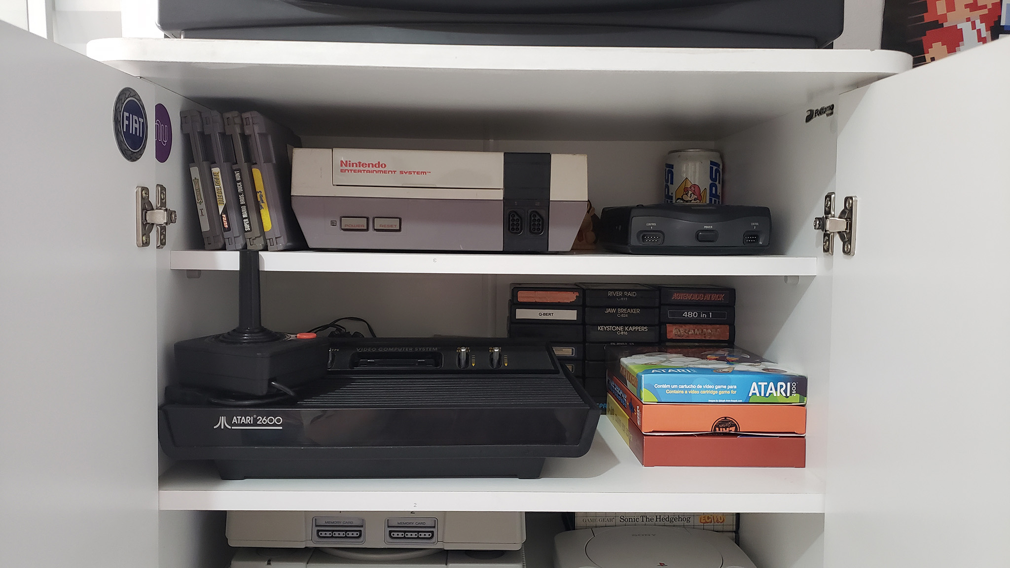 
  
    Na prateleira de cima, o NES e o Sega CDX. Na debaixo, o Atari 2600 com alguns jogos!
  
